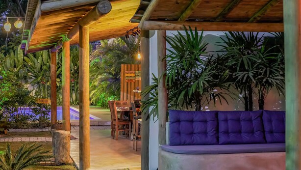 A covered outdoor seating area with a cushioned bench, surrounded by lush tropical plants and a wooden pergola structure.