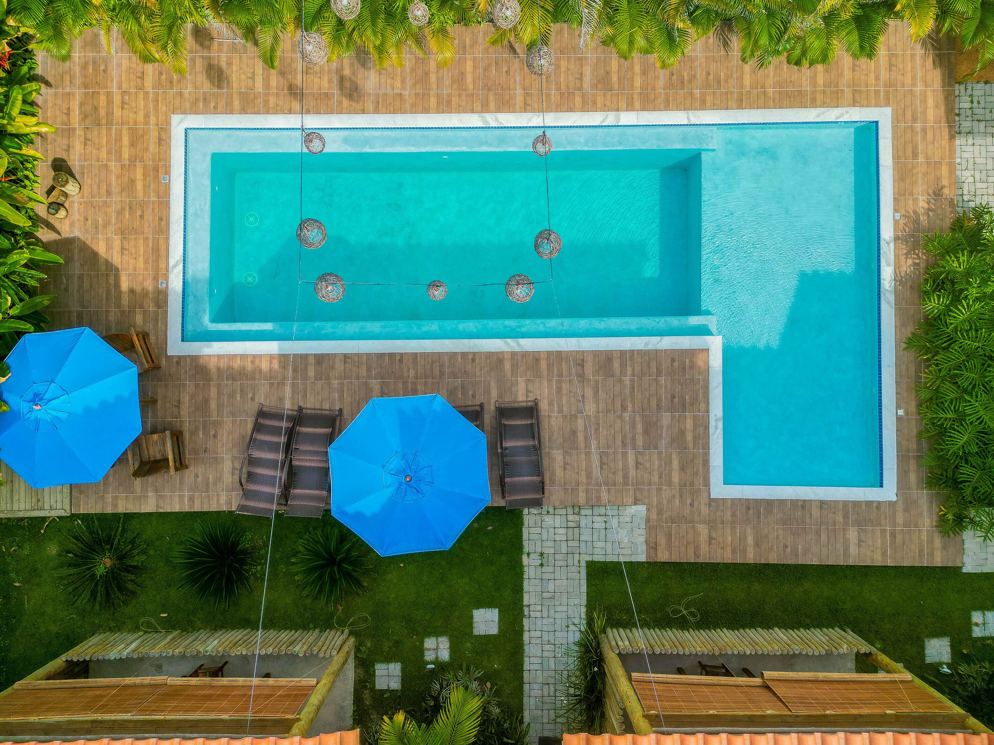 Vue aérienne d’une piscine rectangulaire entourée d’une terrasse en bois, aménagée avec des chaises longues et des parasols.