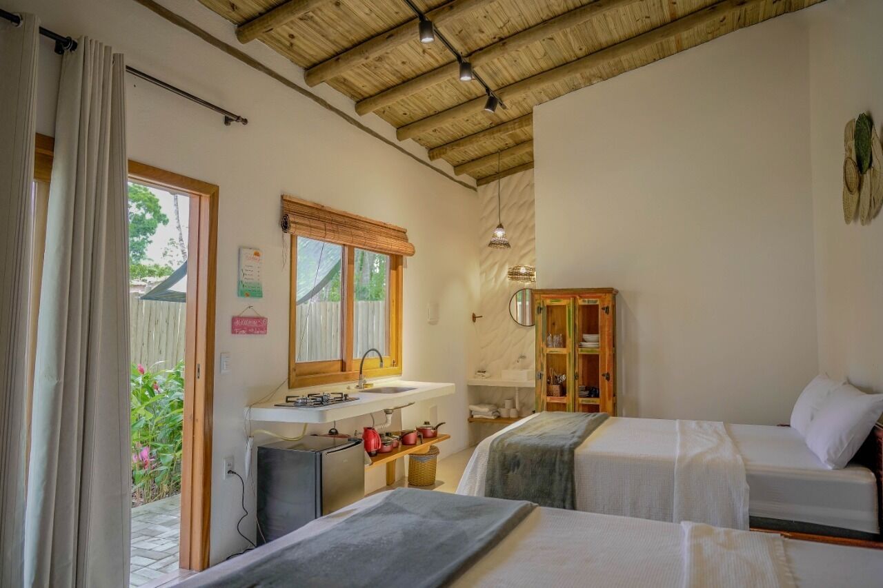 A bedroom with two beds, a wooden ceiling, a sink, and a window with a view of greenery.