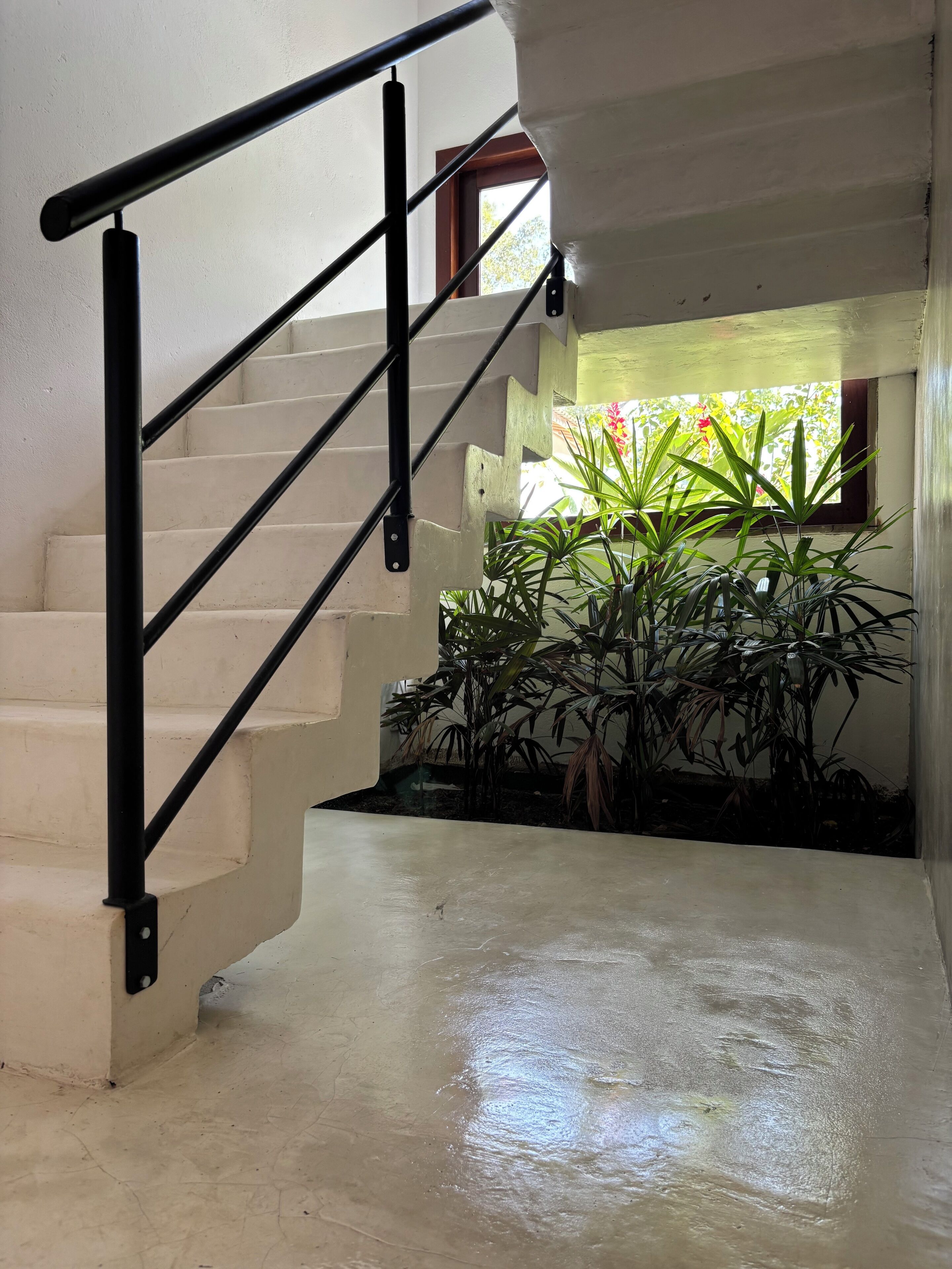 Un escalier avec une rampe en métal noir, des murs de couleur beige et une fenêtre ornée de plantes.