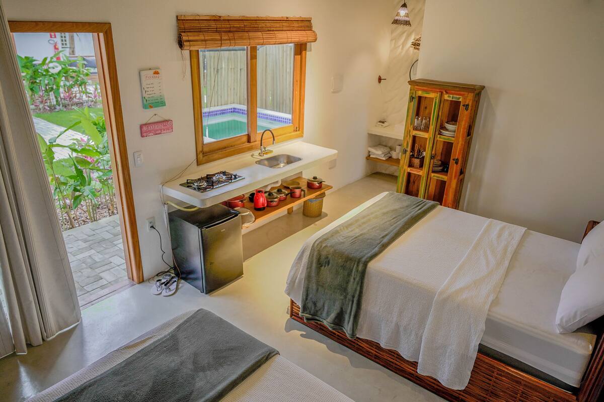 A bedroom with a bed, a small kitchenette, a wooden cabinet, and a view of a garden through a door.