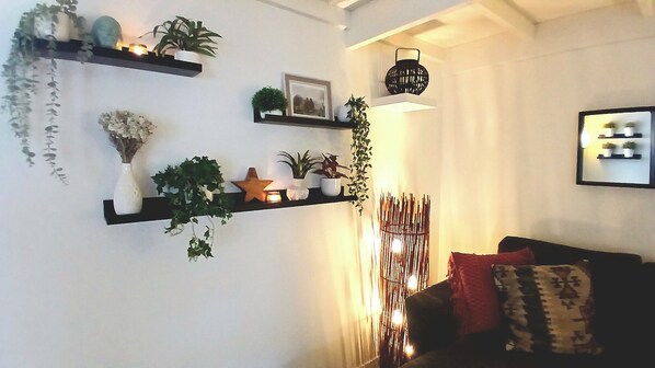 Uma sala de estar com sofá, prateleira fixada na parede com plantas, um quadro emoldurado e um objeto decorativo na parede.