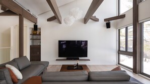 Uma sala de estar moderna com uma televisão de tela grande, um sofá modular cinza, vigas de madeira e um janelão.