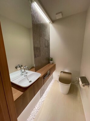 A modern bathroom with a wooden vanity, a white sink, a large mirror, and a wall-mounted toilet. - Silver Maple Chalet (Hakuba)