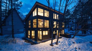 Uma casa moderna, de vários andares, com janelas amplas, cercada por neve e árvores.
