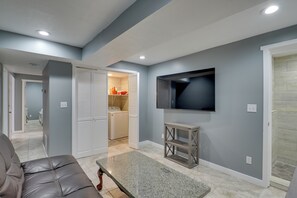 Un salon avec un canapé en cuir, un téléviseur à écran plat fixé au mur et un coin buanderie équipé d’un lave-linge et d’un sèche-linge.