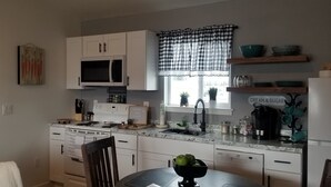 Una cocina con gabinetes blancos, microondas, estufa, lavamanos y ventana con cortina a cuadros.