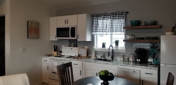 A kitchen with white cabinets, a microwave, a stove, a sink, and a window with a checkered curtain. - Soak up the mountain View in " R Agnew Cottage" (Port Angeles)