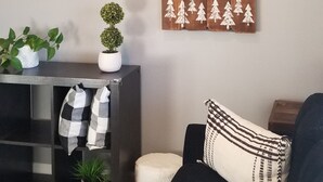 Una sala de estar con un sofá negro, un adorno de pared de madera con un oso y árboles, un televisor de pantalla plana y una mesita con plantas y un mantel a cuadros.