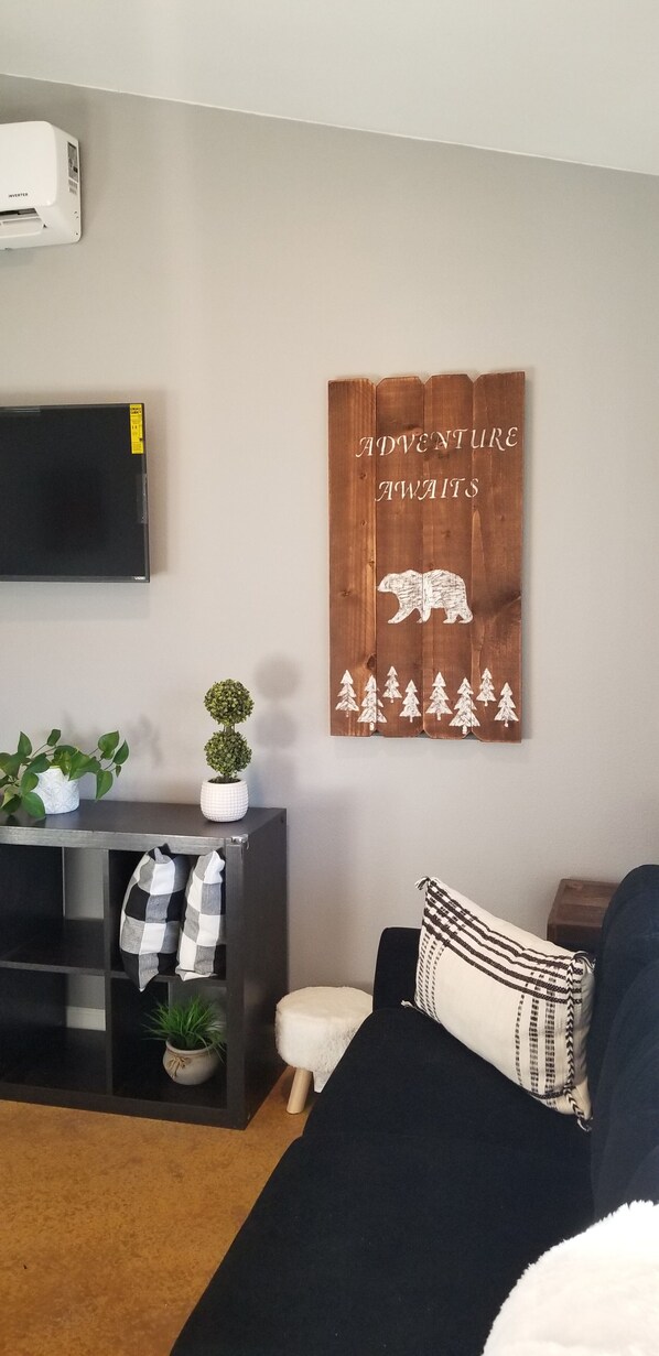A living room with a black sofa, a wooden wall decoration with a bear and trees, a flat-screen TV, and a small table with plants and a checkered cloth. - Soak up the mountain View in " R Agnew Cottage" (Port Angeles)