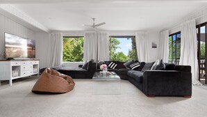 A modern living room with a sectional sofa, a bean bag, a flat-screen TV, and large windows with curtains. - Uralla | Lovely house with pool (West Bathurst)