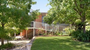 A well-maintained garden with a wooden bridge, a stone planter, and a brick building in the background. - Uralla | Lovely house with pool (West Bathurst)