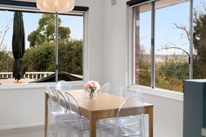 Ein Essbereich mit einem Holztisch und transparenten Stühlen, einer Vase mit Blumen und großen Fenstern mit Blick auf Bäume und einen Zaun.