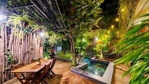 Zona de pati amb taula i caires de fusta, envoltada de vegetació abundant i una petita piscina.
