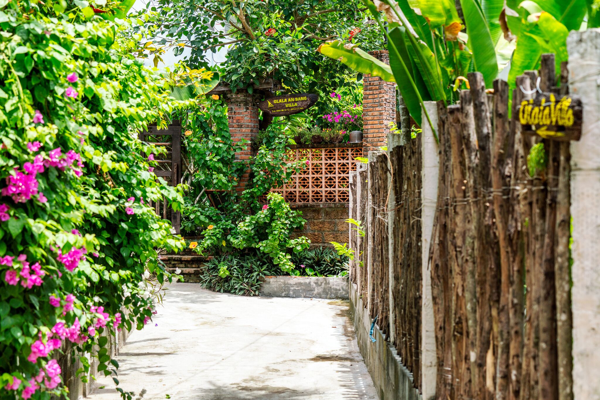 A narrow pathway flanked by lush greenery and a wooden fence, leading to a brick structure with a sign that reads "DOLLA AN BANG VILLA".