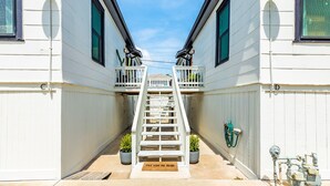 A narrow outdoor corridor with a white staircase leading to a balcony, flanked by white walls and potted plants. - Modern dog-friendly duplex, 1 block from beach with shared backyard patio & W/D (Galveston)