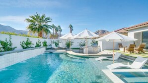 A clear blue pool with lounge chairs and umbrellas, surrounded by a white wall and palm trees. - Pool, fire pit & hot tub in La Quinta (La Quinta)