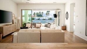 A modern living room with a large flat-screen TV, a sofa, and a wooden coffee table. There is a view of a pool and palm trees through large windows. - Pool, fire pit & hot tub in La Quinta (La Quinta)