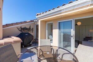 An outdoor patio area with a glass table, chairs, a grill, and a sliding glass door leading to an interior room.