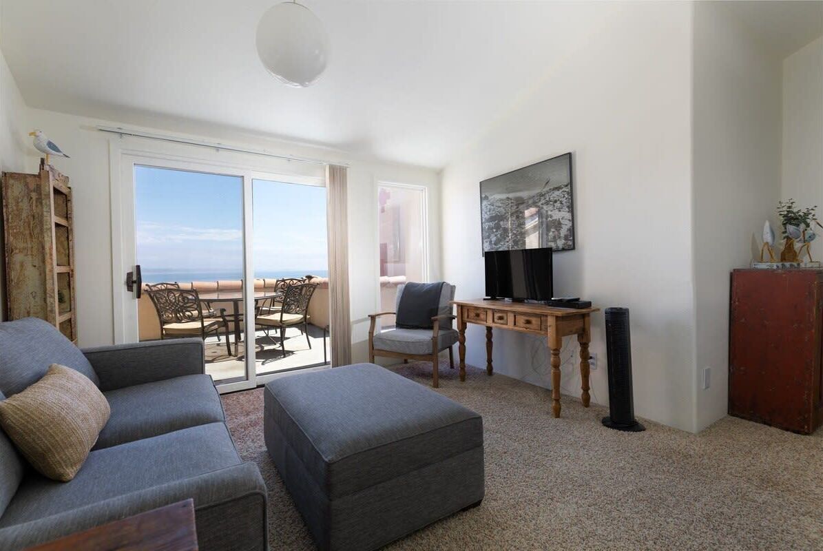 A living room with a sofa, ottoman, and a TV on a wooden stand. There is a sliding glass door leading to an outdoor seating area.