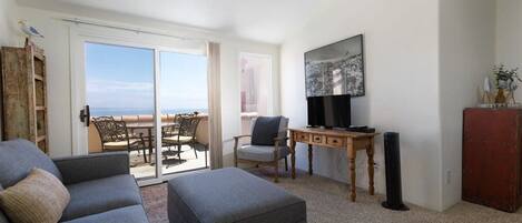 A living room with a sofa, ottoman, and a TV on a wooden stand. There is a sliding glass door leading to an outdoor seating area.