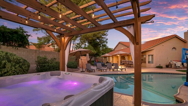 A hot tub under a wooden pergola with a pool and outdoor furniture.