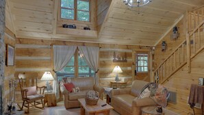 L’intérieur d’une cabane en rondins présente un espace de vie chaleureux, avec des meubles en bois, un fauteuil à bascule et un escalier.