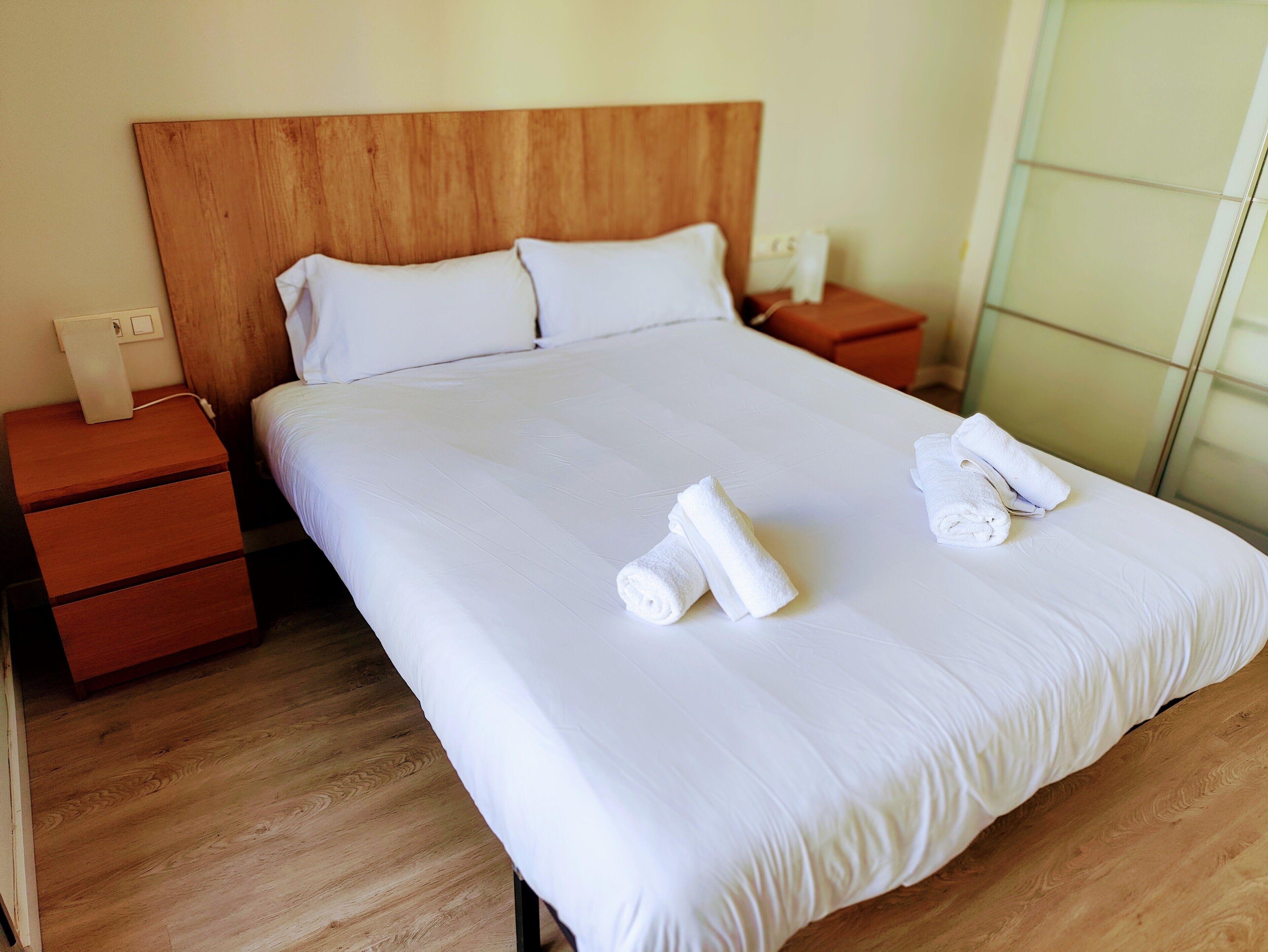 A neatly made bed with white linens and two rolled towels on the bed. There are wooden nightstands with a lamp and a phone on each. The room has wooden flooring and a wooden headboard.