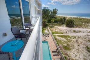 A balcony with wicker chairs and a small table, overlooking a beach with a clear view of the ocean.