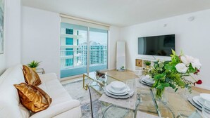 A modern living room with a glass coffee table, white sofa, and a large TV. - TC-3406 · Lovely 2BR Rental in the Heart of Brickell (Miami)