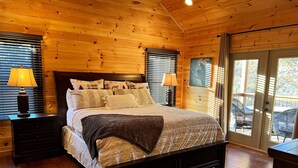 A wooden-paneled bedroom with a large bed, two bedside tables with lamps, and a view through a glass door. - Hot tub, firepit, outdoor living, comfy beds, and more! - 5 O'Clock Somewhere (Mineral Bluff)
