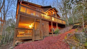A wooden cabin with a balcony, surrounded by trees and a forest floor covered with red mulch. - Cozy Charm with a Fireplace Feature - American Dream (Blue Ridge)