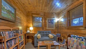 Ein gemütliches Blockhaus-Interieur mit Holzboden, einem grauen Sofa, einem kleinen Tisch, einer Bücherwand und einem gesteppten Sessel.