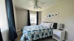 A bedroom with a bed, a nightstand, a ceiling fan, a window with curtains, and framed pictures on the wall.
