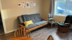 A cozy living room with a grey sofa, wooden chairs, a small table, and a window with a view of a car outside.