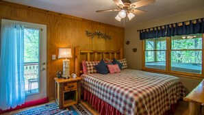 A cozy bedroom with a wooden headboard, a checkered bedspread, a nightstand with a lamp, and a window with a view of greenery.