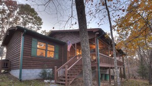 A wooden cabin with a staircase leading to a deck, surrounded by trees and a stone pathway.