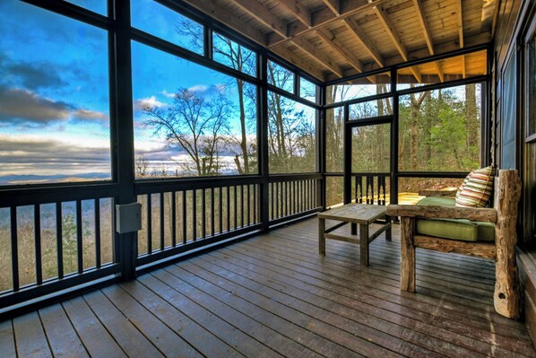 A covered wooden deck with a bench and table, large windows offering a view of trees and a body of water.