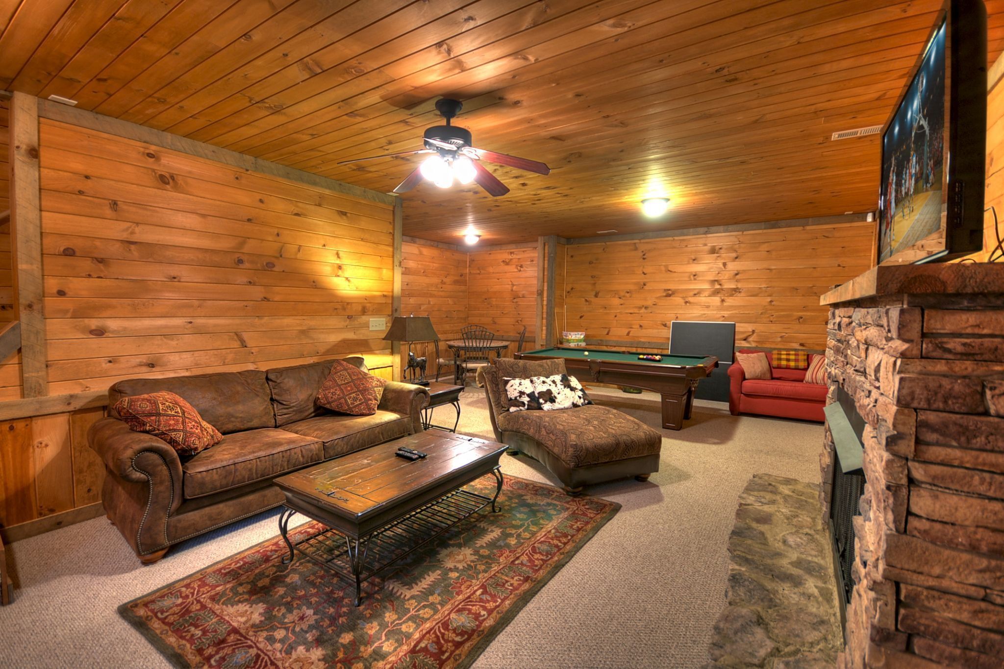 A cozy cabin interior with wooden walls, a ceiling fan, a pool table, a red sofa, and a fireplace.