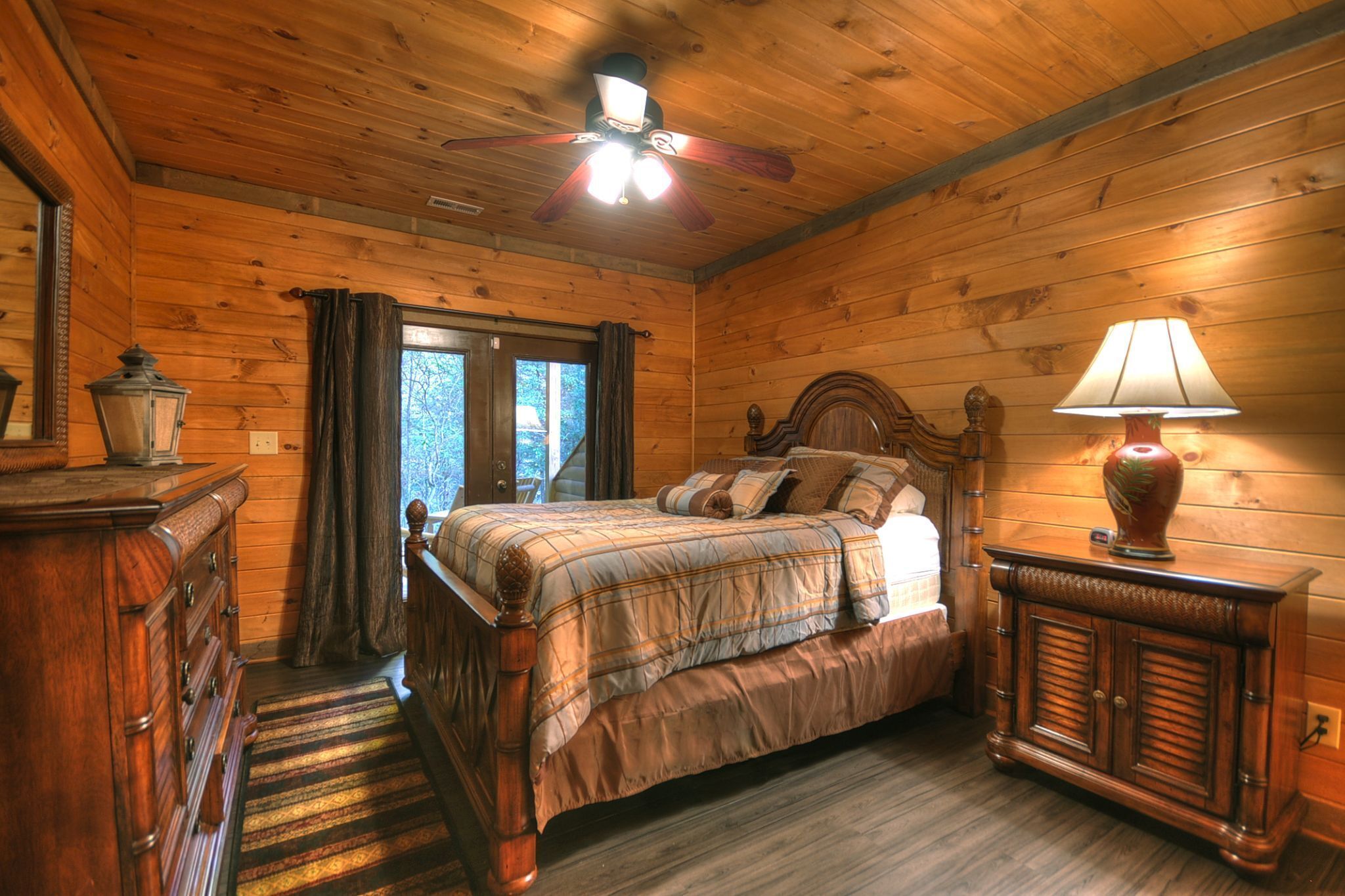 A wooden cabin bedroom with a bed, dresser, lamp, and a ceiling fan.