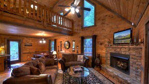 A cozy cabin interior with a fireplace, leather sofas, a dining area, and a large window overlooking a forest. - Nearby creek, cozy hot tub, pool table, and more! - Castle Creek (Cherry Log)