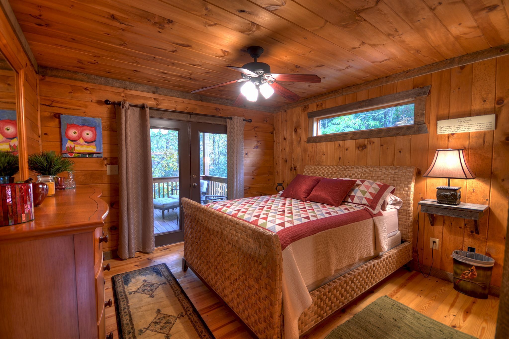 A wooden cabin bedroom with a bed, a ceiling fan, a window, a lamp, and a small table.