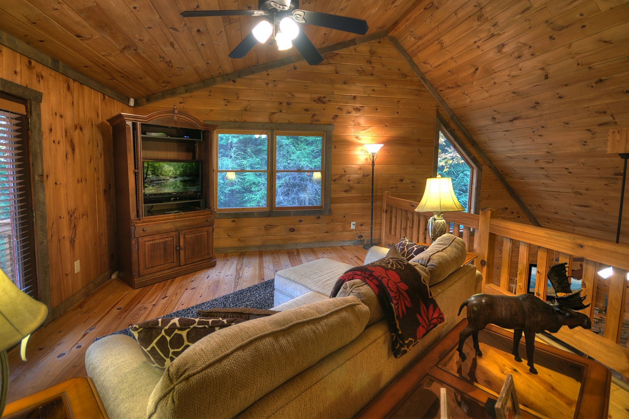 A cozy cabin interior with a wooden ceiling, a television, a sofa, a lamp, and a moose statue.