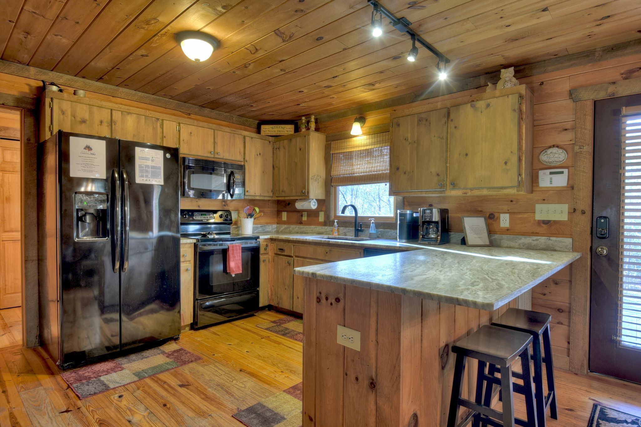 Cocina de cabaña de madera con refrigerador, microondas y estufa de acero inoxidable. Hay una isla de madera con taburetes y una ventana con persianas.