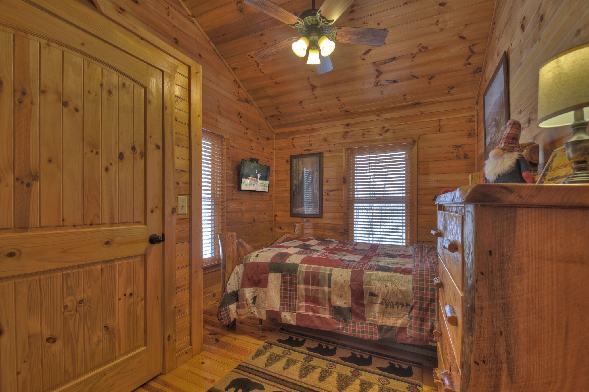 A wooden cabin bedroom with a bed, a dresser, a ceiling fan, and a window with blinds.