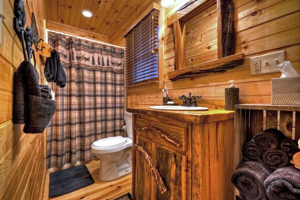 A wooden cabin bathroom with a plaid shower curtain, a white toilet, a wooden sink vanity, and towels stacked on the right.