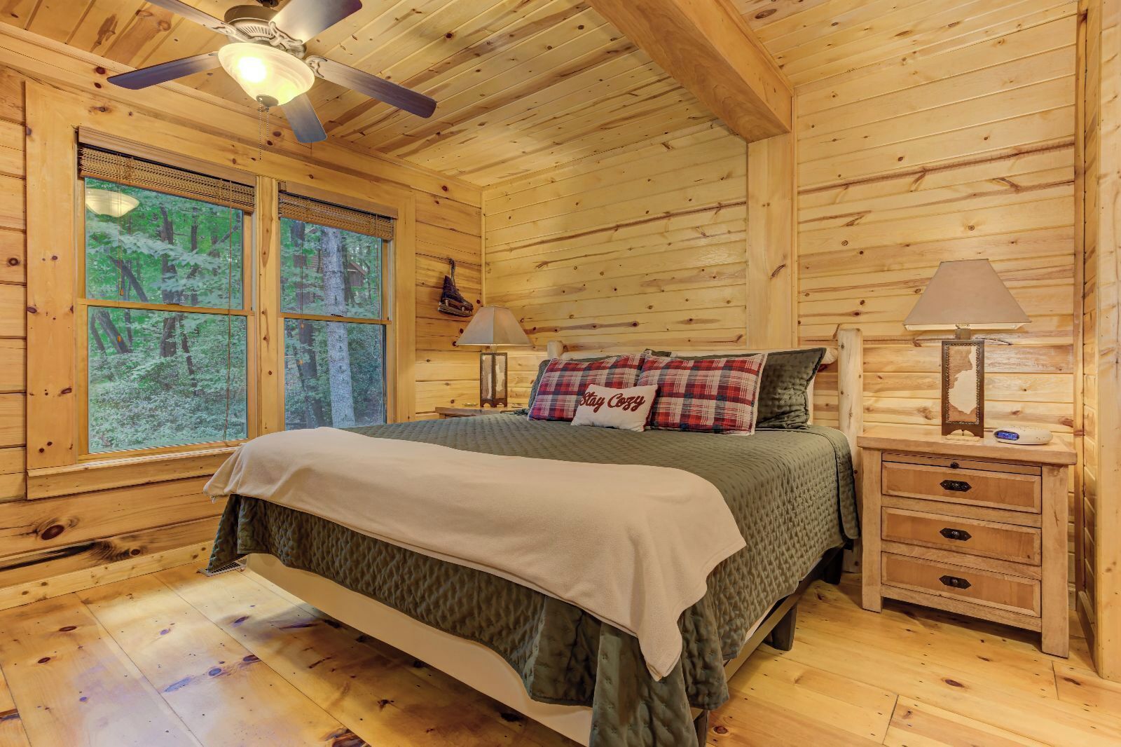 Une chambre située dans une cabane en bois, comprenant un lit, deux tables de chevet, un ventilateur de plafond et de grandes fenêtres donnant sur la forêt.