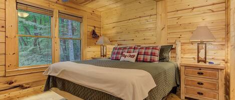 Une chambre située dans une cabane en bois, comprenant un lit, deux tables de chevet, un ventilateur de plafond et de grandes fenêtres donnant sur la forêt.