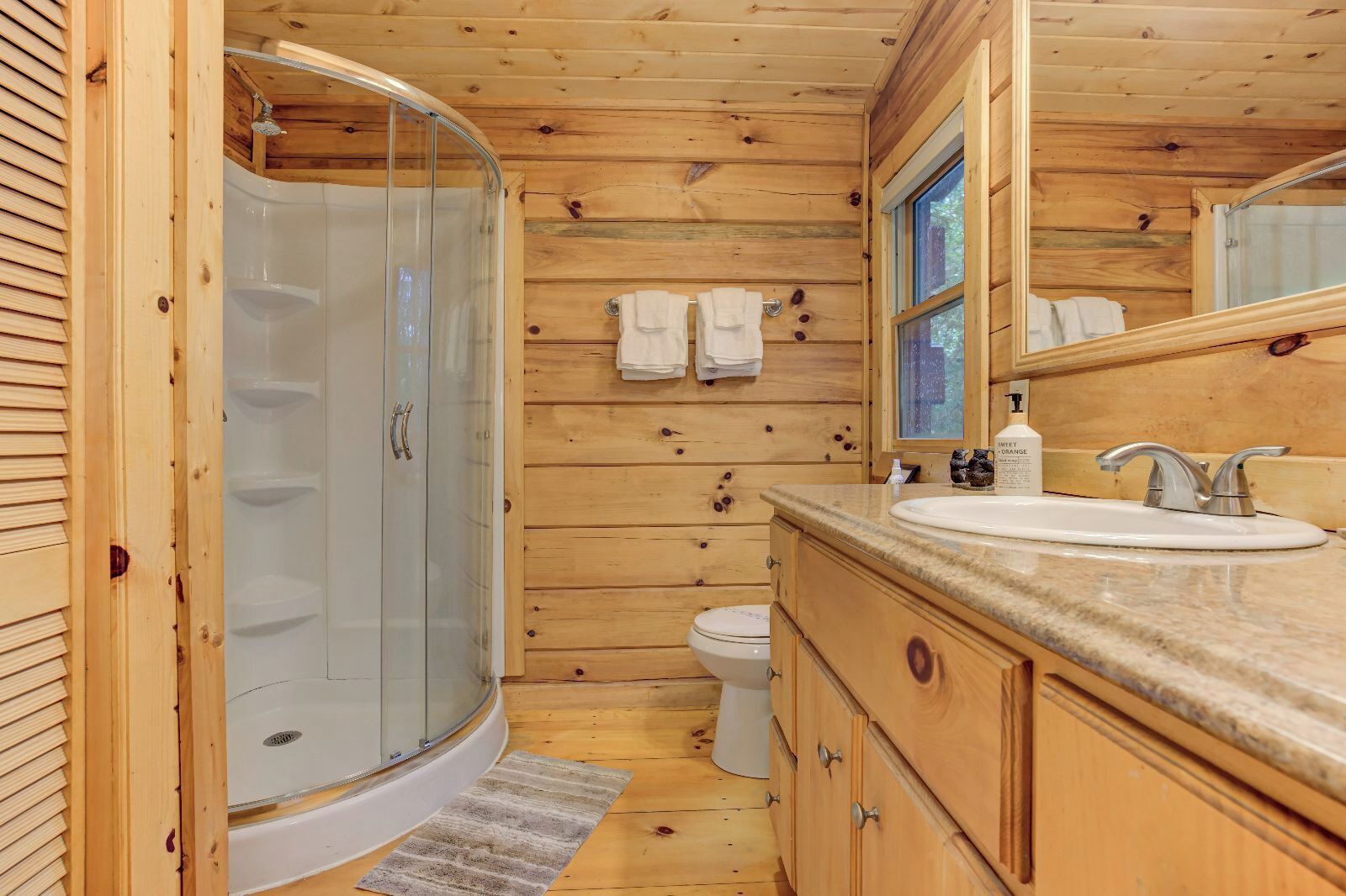 A wooden cabin bathroom with a glass-enclosed shower, a toilet, a sink with a countertop, and wooden cabinets.