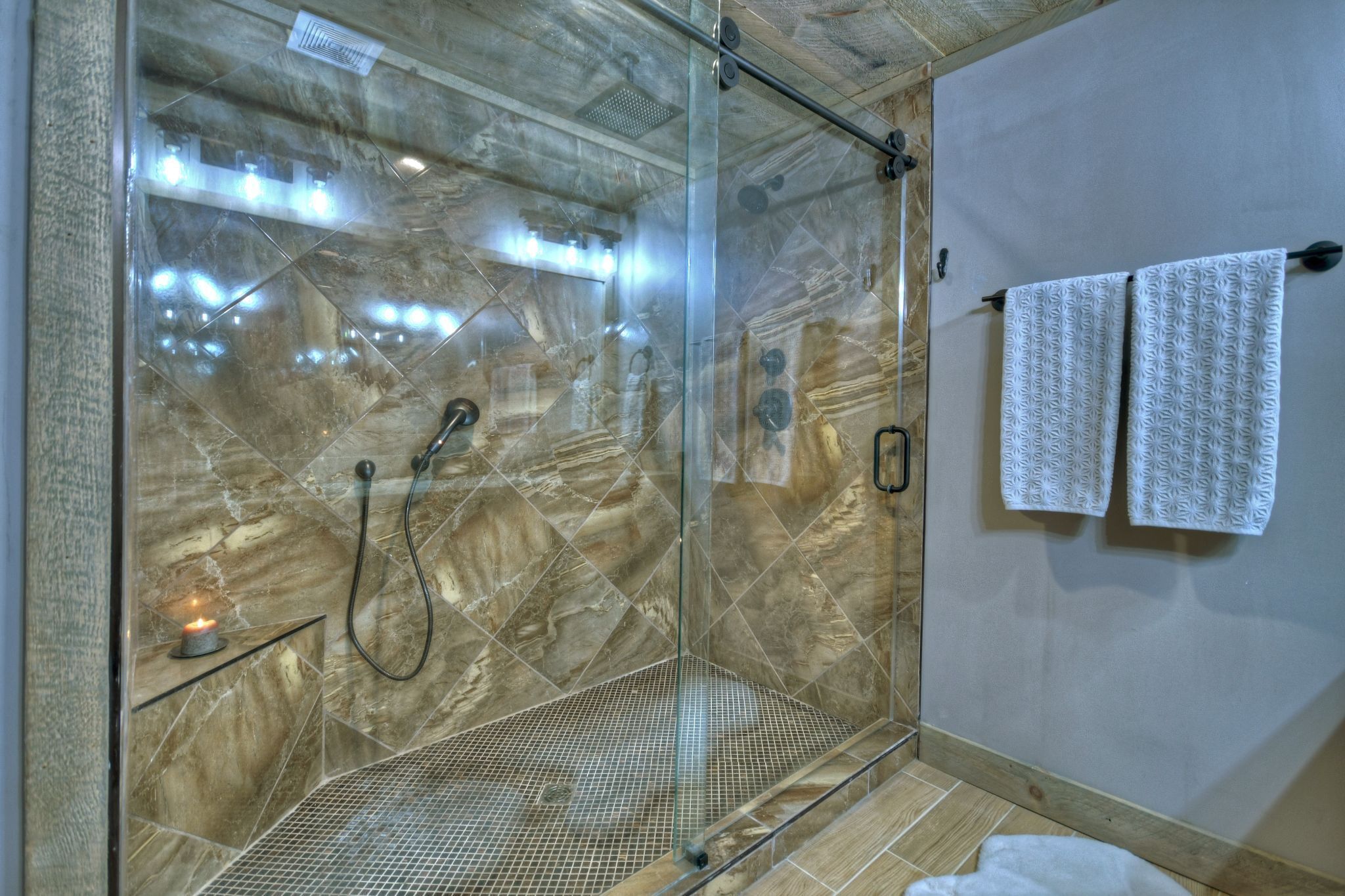 Una ducha con cerramiento de vidrio, pared de mármol, un perchero con dos toallas y una vela pequeña sobre una repisa.